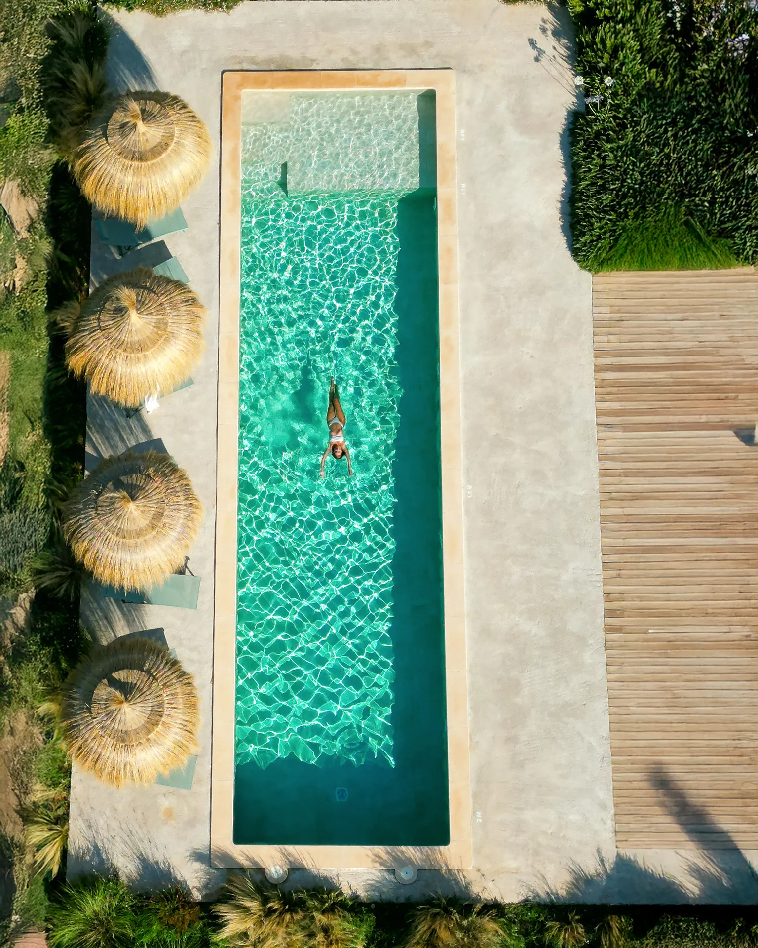 The pool from above — a swimmer cutting through turquoise water, parasols casting shadows